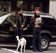 a man and woman standing in front of a black suv with a white dog on the street