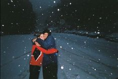 two people standing in the snow hugging each other and smiling at the camera while it's snowing