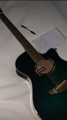 an acoustic guitar sitting on top of a white sheet covered bed next to a notepad