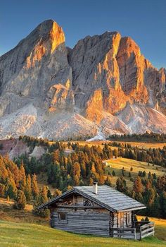 an old cabin sits in front of the mountain range at sunset, with autumn colors on the trees and grass around it