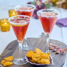 two glasses filled with drinks sitting on top of a wooden platter next to chips