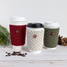 three coffee cups with crochet designs on them sitting next to some pine cones