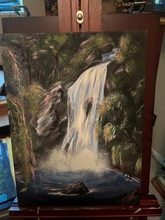 an easel with a painting of a waterfall in the middle and trees around it