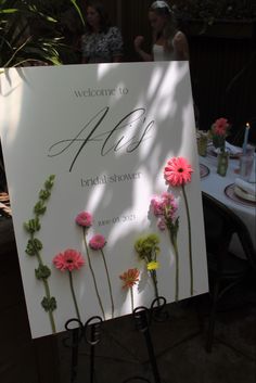 a sign with flowers on it sitting in front of a table and people standing around