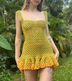 a woman wearing a yellow crochet dress and red earrings standing in the grass