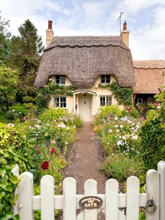 a white picket fence in front of a thatched roof house with flowers around it