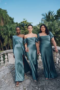 three women in long dresses standing on a stone bridge with palm trees behind them and one woman holding a flower