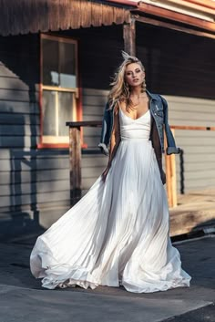 a woman in a white dress and jean jacket standing on the street with her hands behind her back