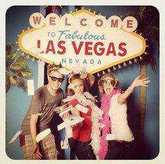 three people posing in front of the welcome sign for las vegas, with palm trees and lights behind them