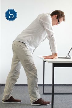 a man standing at a desk with his foot on the table and looking down while using a laptop computer