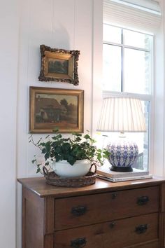 a wooden dresser topped with a potted plant next to a lamp and painting on the wall