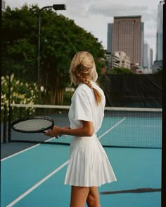 a woman holding a tennis racquet on top of a tennis court with buildings in the background