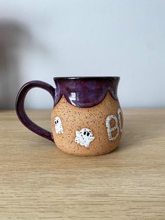 a ceramic mug with ghost faces painted on the side and purple rim sitting on a wooden table