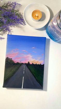 a table with a candle, plate and vase on it that has a painting of a road going into the distance