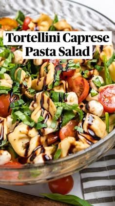 a glass bowl filled with pasta salad on top of a wooden table next to tomatoes