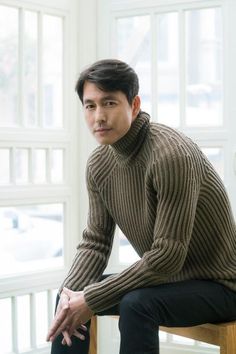 a man sitting on top of a wooden chair wearing a turtle neck sweater and black pants