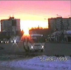 the sun is setting over some buildings and cars in front of an apartment building with snow on the ground