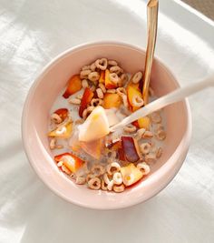 a pink bowl filled with cereal and fruit on top of a white table cloth next to a wooden spoon