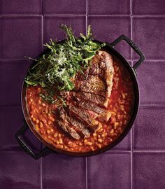 a pan filled with meat and beans on top of a purple tiled countertop next to a green leafy garnish