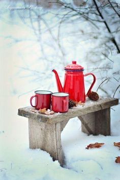 two red mugs are sitting on a bench in the snow with a teapot