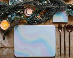 a place setting with silverware, candles and greenery on a wooden table top