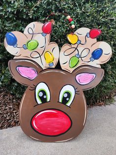 a cardboard reindeer head with christmas lights on it's antlers and nose is sitting in front of a bush