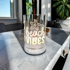 a glass with the words beach vibes on it sitting on a table in front of a potted plant