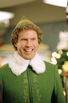 a man wearing a green elf costume with white fur collar smiling at the camera while standing in front of a christmas tree