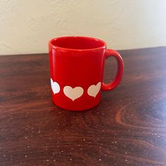 a red coffee mug with white hearts on it sitting on a wooden table next to a wall