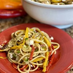 a red plate topped with pasta and vegetables next to a bowl of fettuccine