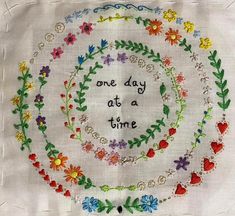 a white cloth with embroidered words and flowers in the center that says, one day at a time