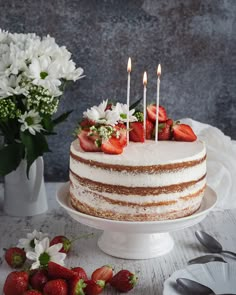 a white cake with strawberries on top and flowers in the background next to it