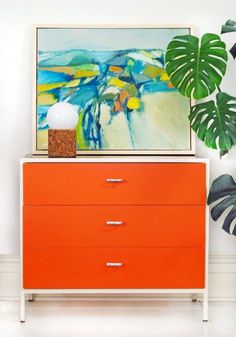 an orange chest of drawers with a painting above it and a potted plant on top