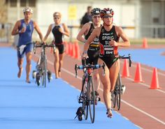 two women racing bikes on a track with orange cones in the foreground and people running behind them