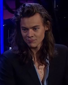 a man with long hair wearing a black suit and blue shirt sitting in front of a tv screen