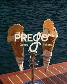 two ice cream cones sitting on top of a wooden table next to the water with words prego gelato italian