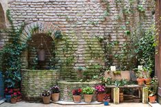 an outdoor fountain surrounded by potted plants in front of a brick wall and doorway