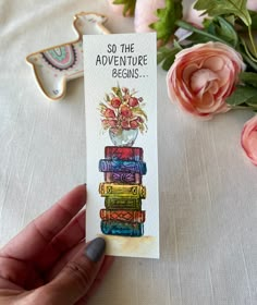 a hand holding a card that says so the adventure begins with flowers and books on it