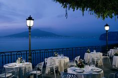 an outdoor dining area with tables and chairs overlooking the water at dusk, lit by street lamps