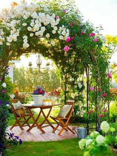 a table and chairs under an arbor in the middle of a garden with flowers on it