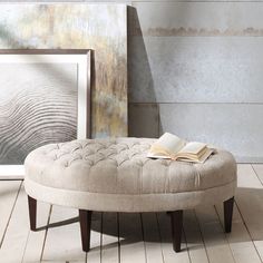 a round ottoman sitting on top of a wooden floor next to a painting and an open book