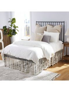 a bed with white sheets and pillows in a room next to a table, plant and window