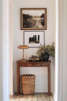 a wooden table topped with a lamp next to a painting and a basket filled with flowers