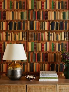 a lamp and some books on a table in front of a book shelf with many books