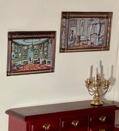 two paintings hang on the wall above a red dresser with candles and a candelabra