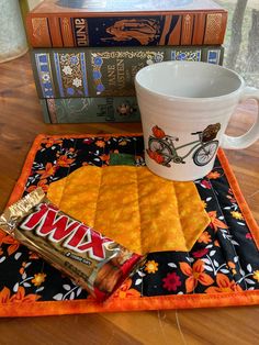 a coffee cup, candy bar and books on a wooden table with an orange place mat