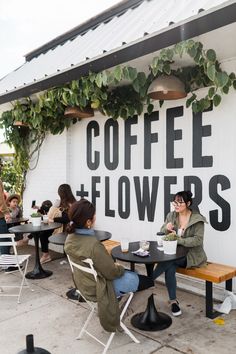 people sitting at tables in front of a coffee and flowers sign on the side of a building