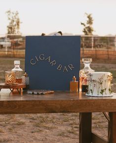 a wooden table topped with bottles and cakes on top of a dirt field next to a fence