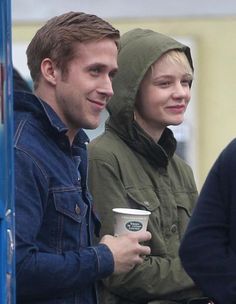 two people standing next to each other holding coffee cups and looking at something in the distance