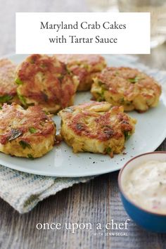 several crab cakes on a plate next to a bowl of cream cheese and a glass of water